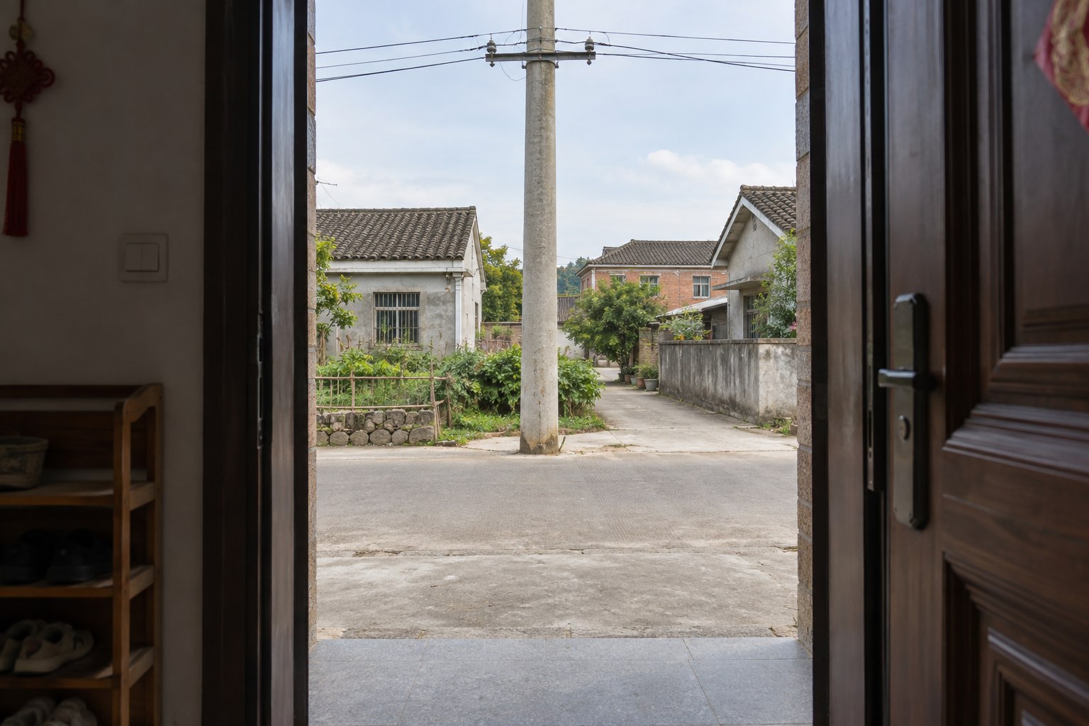 站在敞开的住宅门内,近处一根水泥电线杆位于门框中线,横向村道从门前经过。